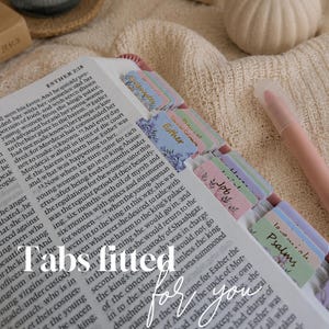 以下が含まれることがあります： 印刷されたテキストが記載された開いた本と、ページの端に取り付けられたカラフルで装飾的な粘着タブのセット。タブには花柄と「Esther」、「Job」、「Psalms」などのラベルがあり、横には淡いピンク色のペンが添えられています。
