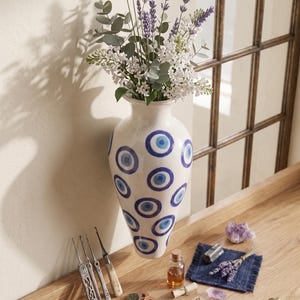 May include: A white vase with blue eye designs holds a bouquet of lavender and white flowers. Various objects are arranged on a wooden surface, including tools, coins, a pen, matches, and glasses. A decorative and artistic display.