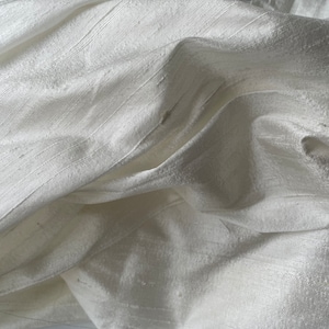 May include: Close-up of a white silk fabric with a smooth, lustrous texture. The fabric is draped and folded, showcasing its soft and flowing nature.