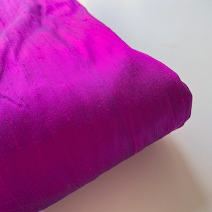 May include: A close-up of a vibrant magenta silk fabric with a subtle sheen. The fabric is folded and has a soft, drapey texture.