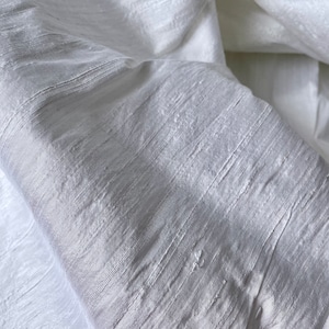 May include: White silk fabric with a subtle, textured, woven pattern. The fabric is folded and draped, showing the soft, flowing texture.