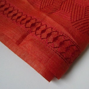 May include: A close-up of a rust-colored fabric with a geometric pattern embroidered in a darker red thread. The fabric has a textured surface and a decorative border.