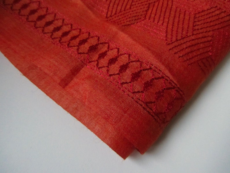 May include: A close-up of a rust-colored fabric with a geometric pattern embroidered in a darker red thread. The fabric has a textured surface and a decorative border.