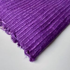 May include: Purple fabric with a textured, ribbed surface. The fabric has a raw, unfinished edge.