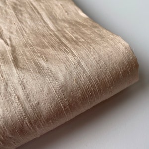 May include: A close-up of a beige silk fabric with a subtle sheen. The fabric has a textured, woven appearance.