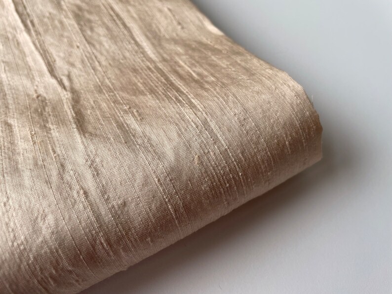 May include: A close-up of a beige silk fabric with a subtle sheen. The fabric has a textured, woven appearance.