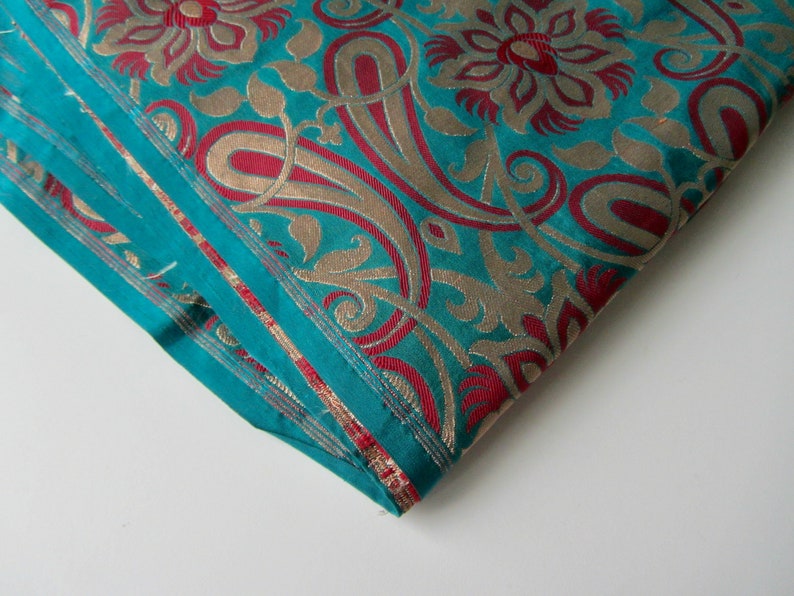K&ouml;nnte beinhalten: T&uuml;rkisfarbener Stoff mit einem goldenen und burgunderroten Paisley-Muster. Der Stoff ist in der Mitte gefaltet und die Kanten sind sichtbar.