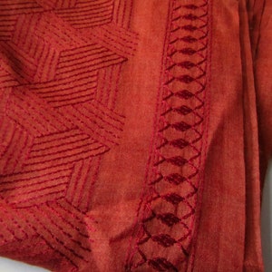 May include: A close-up of a rust-colored fabric with a geometric pattern embroidered in a darker shade of rust. The fabric has a soft, textured feel.