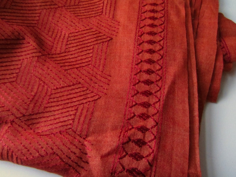 May include: A close-up of a rust-colored fabric with a geometric pattern embroidered in a darker shade of rust. The fabric has a soft, textured feel.