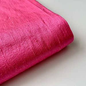 Puede incluir: Primer plano de una tela de color rosa brillante con una textura suave y sedosa. La tela está doblada, revelando el brillo del material.