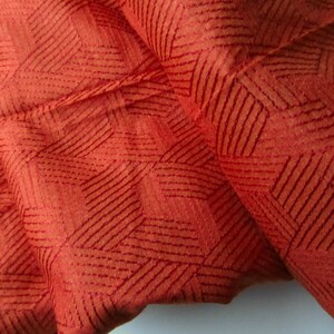 May include: Close-up of a rust-colored fabric with a geometric pattern of thin, dark red lines. The fabric has a textured surface.