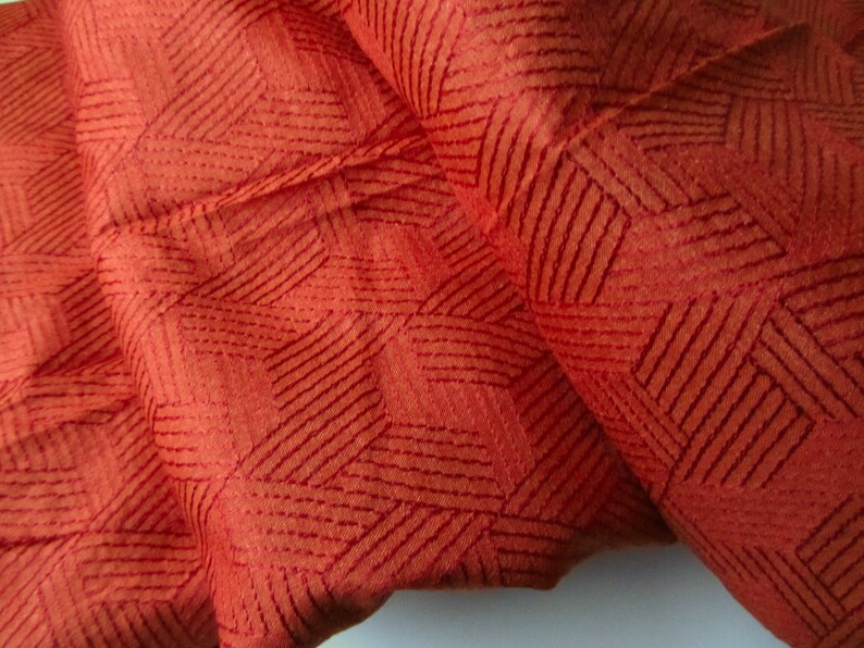 May include: Close-up of a rust-colored fabric with a geometric pattern of thin, dark red lines. The fabric has a textured surface.