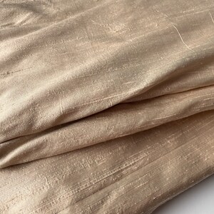 May include: A close-up of a beige fabric with a subtle sheen and a textured, woven pattern. The fabric is folded and draped, showcasing its soft and luxurious feel.