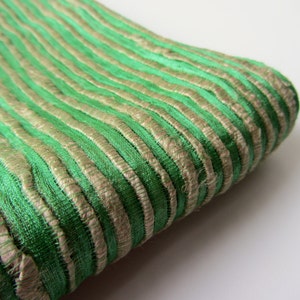 Puede incluir: Una pieza de tela doblada que presenta un patrón repetitivo de rayas verticales alternas en verde vibrante y oro metálico. El material tiene una superficie texturizada y acanalada con un ligero brillo, mostrando un diseño lineal consistente en todo el tejido.