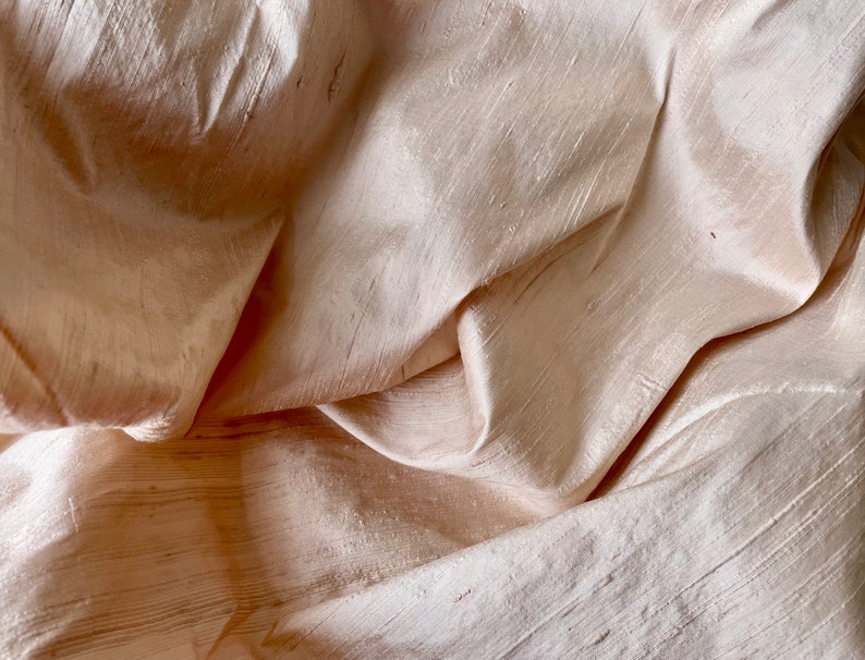 May include: A close-up of a soft, pale peach-colored fabric with a subtle sheen. The fabric is draped and folded, creating a textured surface.