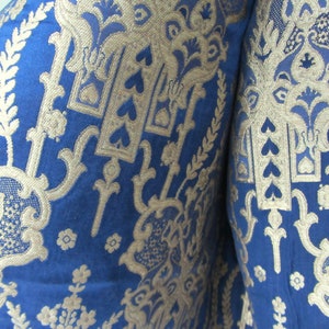May include: Close-up of a blue fabric with a gold pattern. The pattern features intricate floral designs and repeating heart shapes.