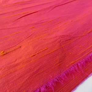 May include: Close-up of a pink fabric with orange stripes. The fabric has a textured, woven look and a pink fringe along the edge.