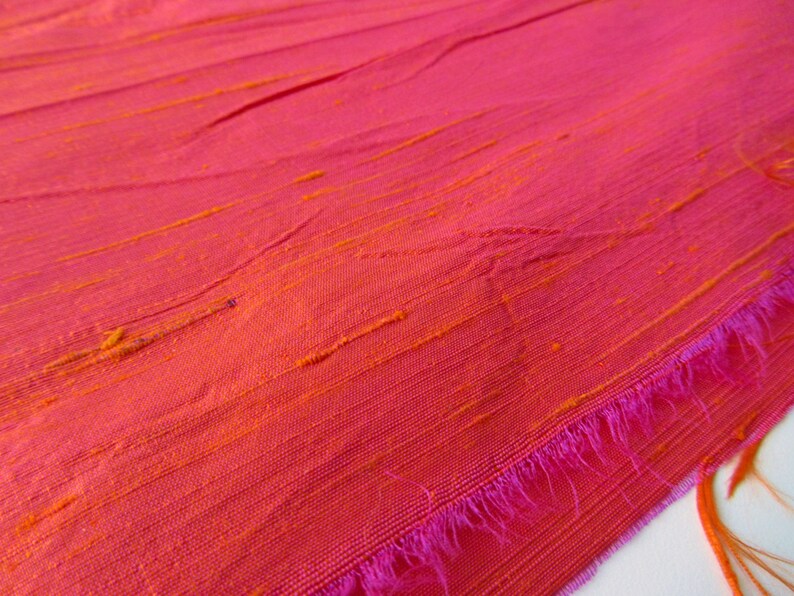 May include: Close-up of a pink fabric with orange stripes. The fabric has a textured, woven look and a pink fringe along the edge.