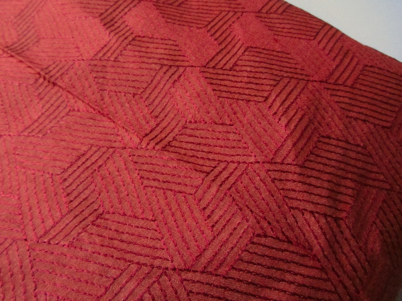 May include: Close-up of a red fabric with a geometric pattern of stitched lines forming a diamond shape.