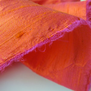 May include: Close-up of a bright orange and pink fabric with a frayed edge. The fabric has a subtle texture and a slightly iridescent sheen.