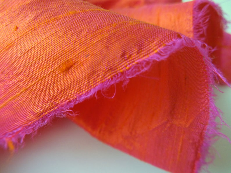 May include: Close-up of a bright orange and pink fabric with a frayed edge. The fabric has a subtle texture and a slightly iridescent sheen.