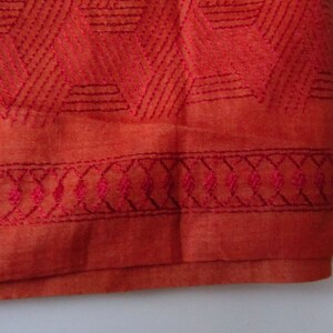 May include: A close-up of the bottom edge of a burnt orange fabric with a geometric pattern embroidered in a darker shade of orange. The fabric has a subtle sheen and a soft texture.