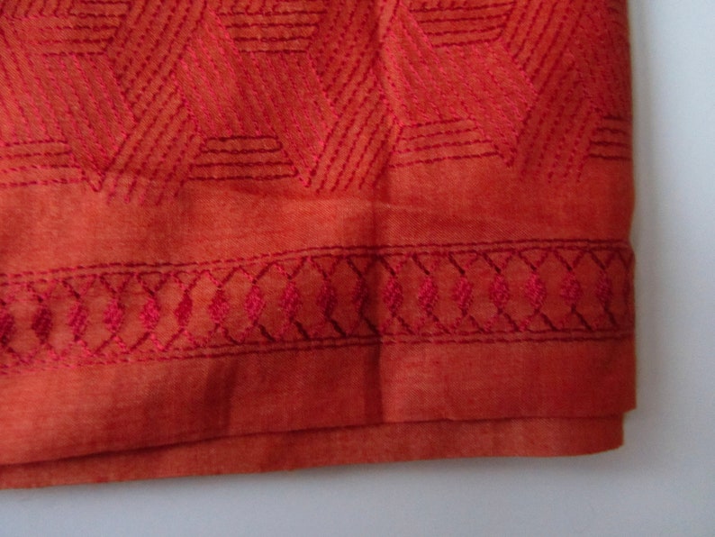 May include: A close-up of the bottom edge of a burnt orange fabric with a geometric pattern embroidered in a darker shade of orange. The fabric has a subtle sheen and a soft texture.