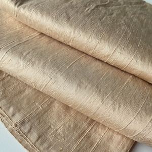 May include: A close-up of a folded piece of beige silk fabric with a subtle sheen. The fabric has a smooth, luxurious texture and a slightly wrinkled appearance.