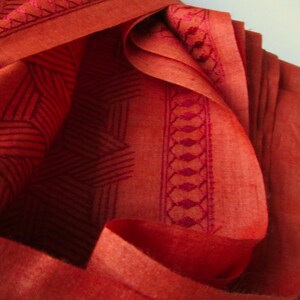 May include: Close-up of a folded piece of fabric with a geometric pattern in a deep red color. The fabric has a subtle sheen and a textured surface.