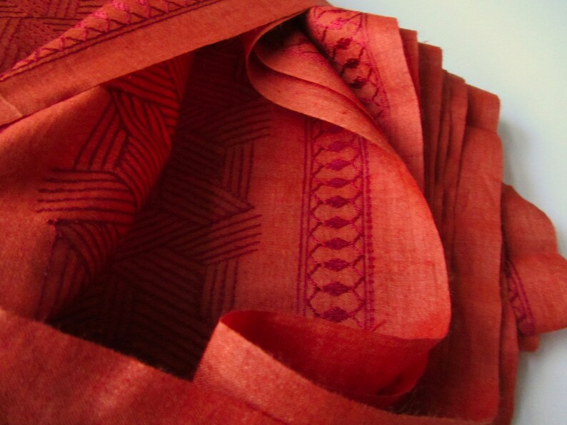 May include: Close-up of a folded piece of fabric with a geometric pattern in a deep red color. The fabric has a subtle sheen and a textured surface.