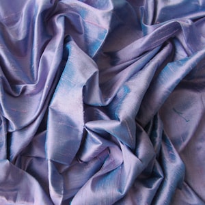 May include: A close-up shot of a draped fabric with a shimmering, iridescent quality. The fabric is primarily a soft lavender color, with hints of blue and pink. The folds and creases create a textured, flowing appearance, suggesting a luxurious material.
