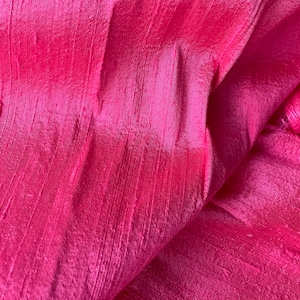 May include: Close-up of a pink silk fabric with a textured, woven pattern. The fabric is folded and draped, showing the soft, flowing texture.