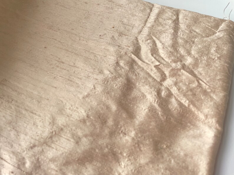 May include: A close-up of a piece of beige silk fabric with a subtle sheen. The fabric has a wrinkled texture and is folded in a way that shows the drape and flow of the material.