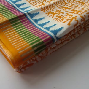 Könnte beinhalten: Ein gefalteter Stoff mit weißem Hintergrund und einem Muster aus braunen stilisierten Figuren und Bäumen. Der Stoff hat einen gelben, grünen, violetten und blauen Rand.