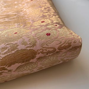 Puede incluir: Primer plano de una tela doblada con un patrón floral detallado. La tela tiene una base rosa pálido con diseños florales dorados y pequeños acentos rojos. La textura parece suave y lujosa.