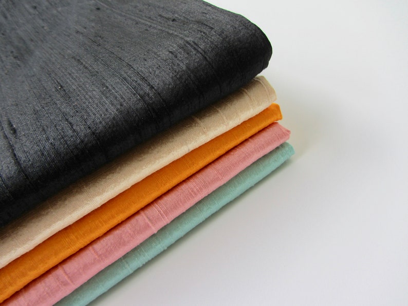 K&ouml;nnte beinhalten: Ein Stapel aus f&uuml;nf Stoffproben in verschiedenen Farben. Die oberste Probe ist schwarz, gefolgt von beige, orange, rosa und hellblau.
