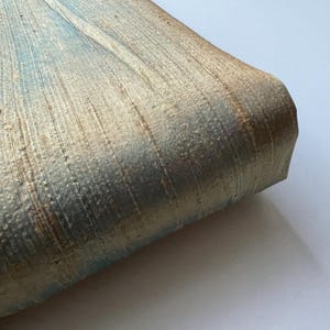 May include: Close-up of a metallic fabric with a subtle shimmer. The fabric has a woven texture with vertical lines and a light blue and gold color scheme.