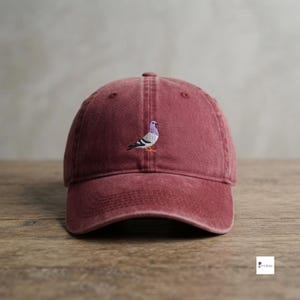May include: A maroon baseball cap with a six-panel construction and a curved brim. The front center of the cap features a small, detailed embroidered design of a pigeon with purple, white, and black feathers and orange feet.