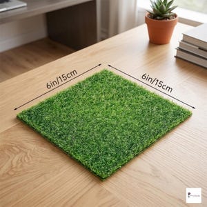 Op de afbeelding: Een vierkant kunstgras, 15 cm per zijde, wordt getoond op een houten oppervlak. Het heldergroene gras contrasteert met het lichtbruine hout. De afmetingen zijn duidelijk aangegeven met zwarte pijlen en tekst.