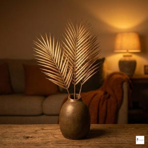 Op de afbeelding: Een donkerbruine, ronde keramische vaas bevat drie metallic gouden palmbladeren. De vaas heeft een gladde, matte afwerking en een smalle hals, terwijl de palmbladeren gedetailleerde, puntige bladeren hebben die in een waaier uitstaan.