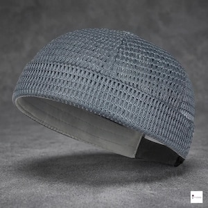 Può includere: Un berretto grigio senza visiera realizzato in tessuto a rete testurizzato a trama aperta. Il cappello presenta una calotta arrotondata con cuciture verticali visibili e una fodera interna in tinta unita chiara lungo il bordo inferiore.