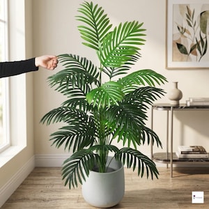 May include: A large, artificial palm plant with vibrant green fronds. The plant is potted in a light gray, modern-style pot. The plant is indoors, and a person's hand is visible, touching the plant's leaves.