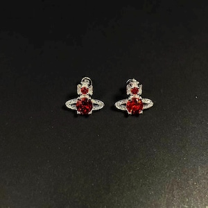 Könnte beinhalten: Ein Paar silberfarbene Ohrstecker mit einem roten herzförmigen Mittelstein. Die Ohrringe haben ein einzigartiges Design mit einem Kranz aus kleinen, klaren Steinen um den roten Mittelstein und einem kleinen roten Stein oben.