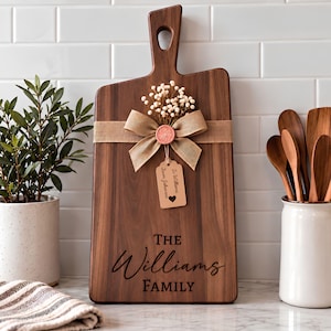 May include: A rectangular dark wood cutting board with a handle featuring a hole. It is decorated with a tan burlap ribbon, a small bouquet of white dried flowers, a pink wax seal, and a tag reading To Williams From Johnsons.