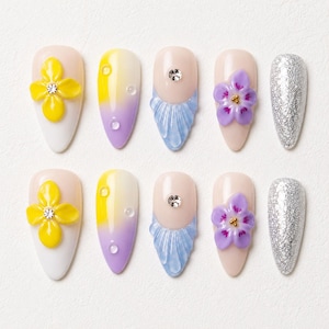 Könnte beinhalten: Ein Set aus zehn mandelförmigen Press-on-Nägeln mit verschiedenen Designs, darunter gelbe 3D-Blumen mit Strasssteinen, ein Gelb-zu-Lila-Ombré mit klaren Tropfen, blaue texturierte Spitzen, lila 3D-Blumen und vollflächiger silberner Glitzer.