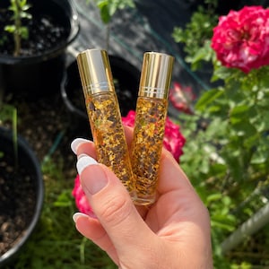 Könnte beinhalten: Zwei transparente Roll-on-Flaschen mit goldenen Kappen werden vor einem verschwommenen Hintergrund aus Pflanzen und Blumen gehalten. Die Flaschen sind mit einer goldenen Flüssigkeit und Goldflocken gefüllt. Die Flaschen werden von einer Person mit weißem Nagellack gehalten.