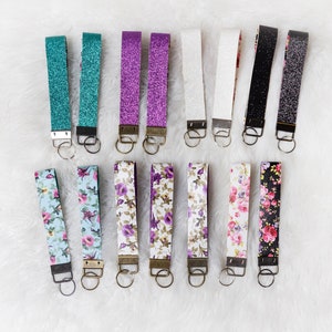 May include: A collection of 14 keychains with different designs. The keychains are made of fabric and have a metal ring. The designs include glitter, floral patterns, and solid colors.