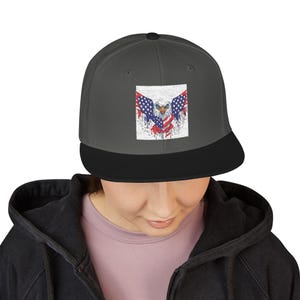 May include: A gray and black baseball cap featuring a distressed graphic of an American bald eagle with the US flag. The eagle's wings are adorned with the stars and stripes, and the cap has a flat brim.