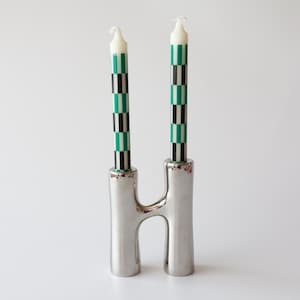 Puede incluir: Un candelabro de metal plateado con silueta en forma de H y dos soportes cilíndricos. Dos velas cónicas están insertadas en los soportes, cada una decorada con un patrón vertical repetitivo de cuadrados alternos en negro, blanco y verde.