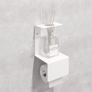 May include: A white wall-mounted metal shelf featuring a top platform with a circular cutout for a clear glass reed diffuser bottle labeled BARRY WHITE GOLD and a lower section designed to hold a standard roll of toilet paper on a horizontal rod.